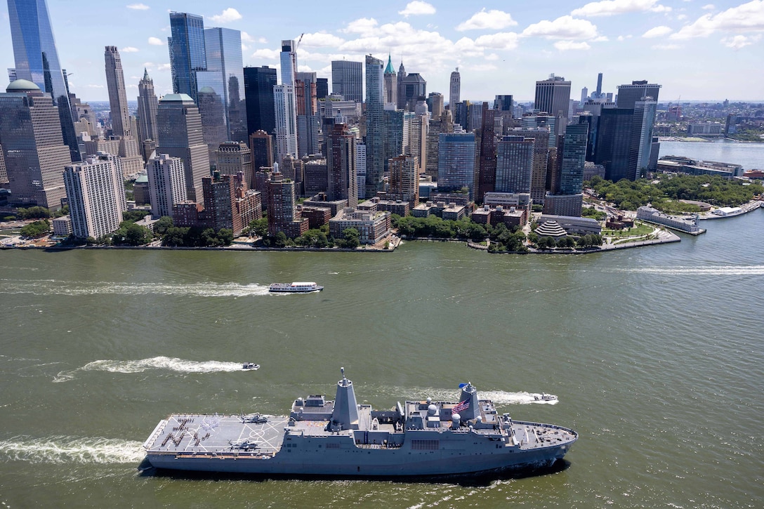 Marines and sailor spell out "I Love New York" on the flight deck of a ship as it transits a body of water with boats near a city's skyline.
