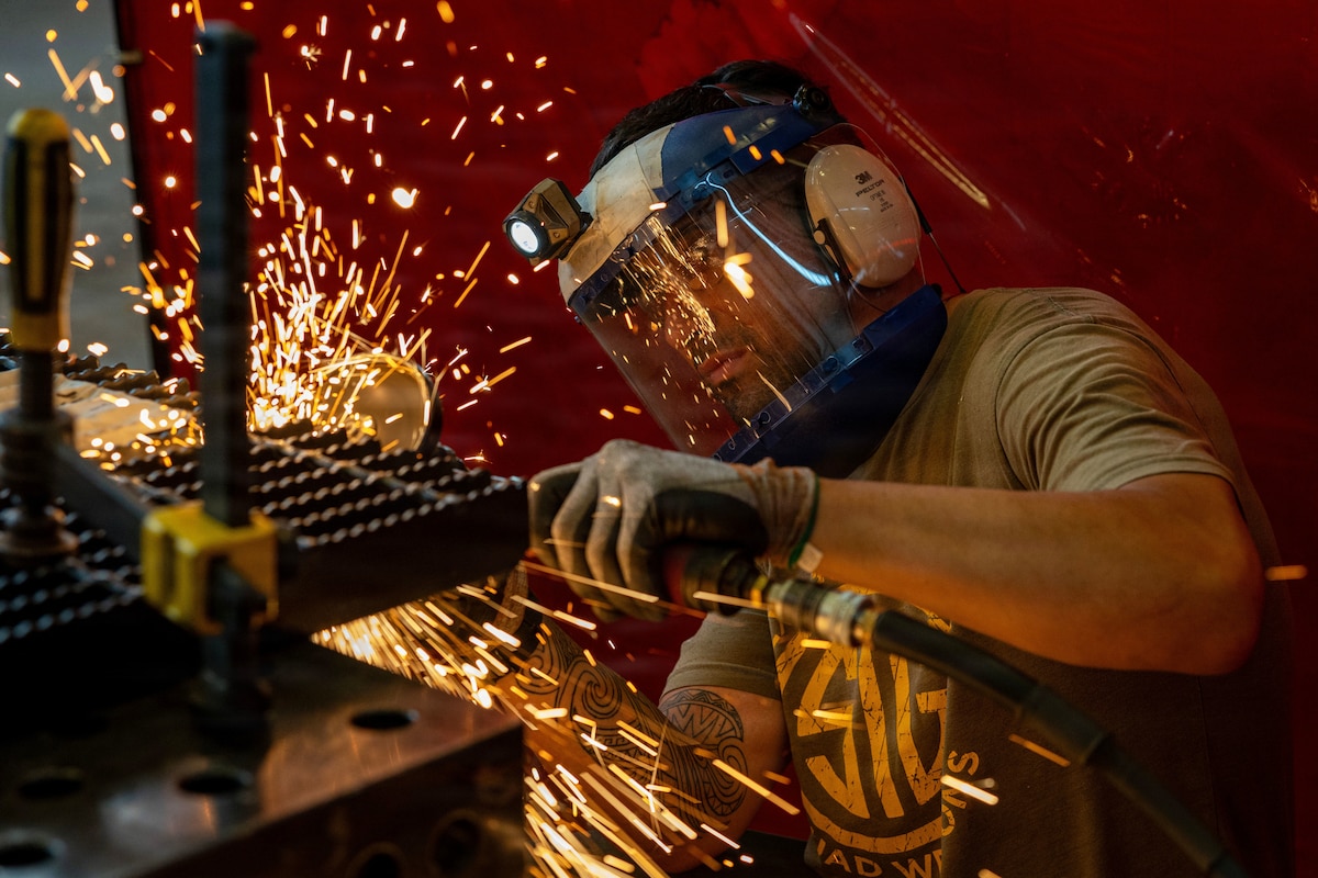 Sparks fly as a shipfitter wearing protective face gear with a headlamp welds metal in a dimly lit room.