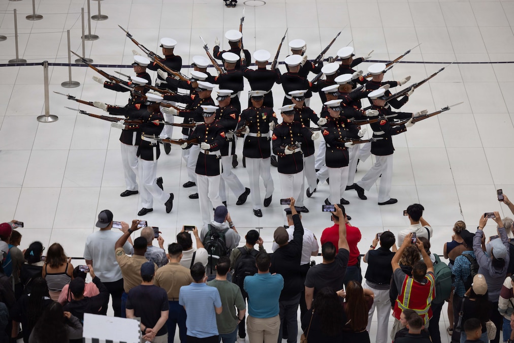 Marines in blue and white uniforms aim rifles in a circular formation as spectators watch and record as seen from above.
