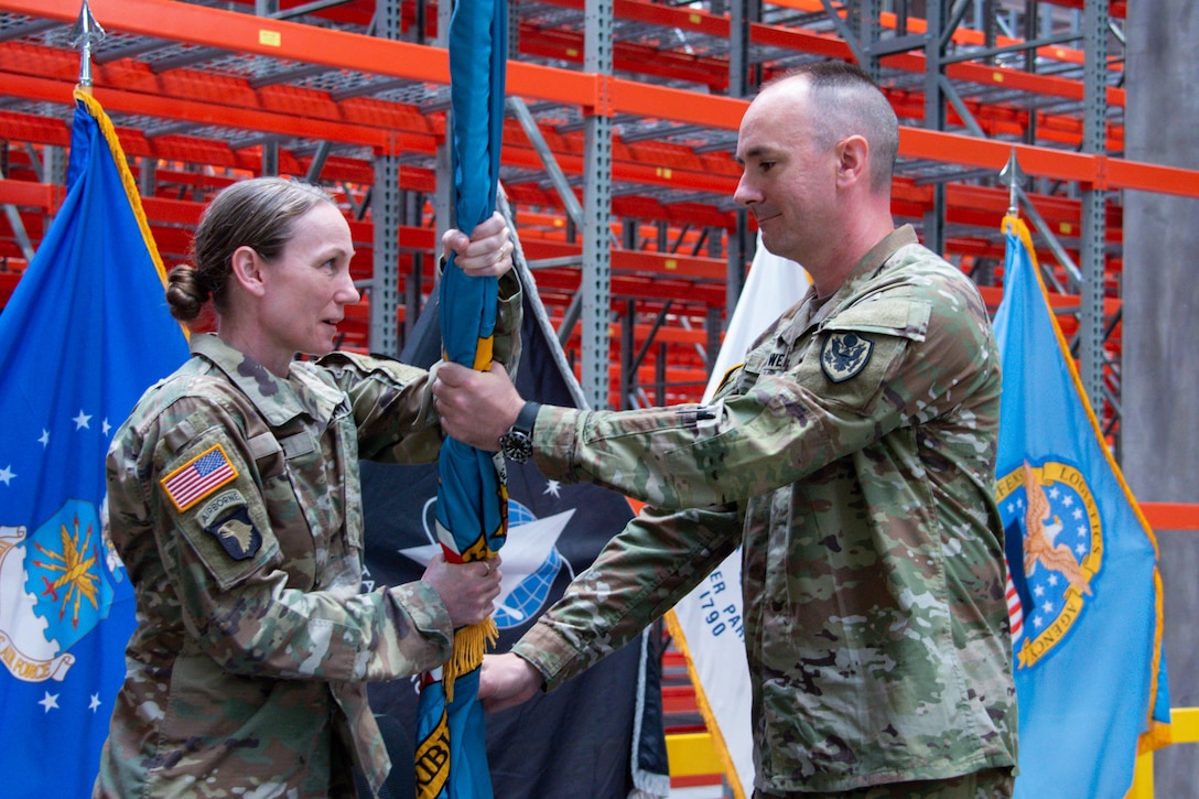 People wearing camouflage military uniforms stands inside a warehouse passing a flag.