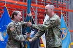 People wearing camouflage military uniforms stands inside a warehouse passing a flag.