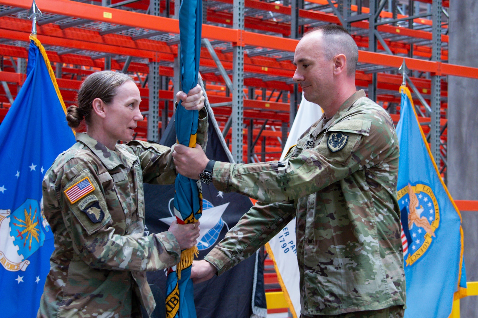 People wearing camouflage military uniforms stands inside a warehouse passing a flag.