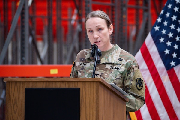 A woman wearing a camouflage military uniform stands inside a warehouse behind a podium speaking.