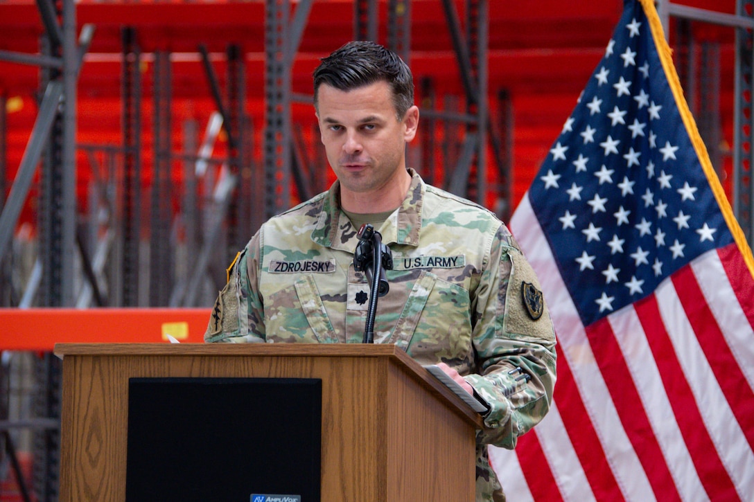 A man wearing a camouflage military uniform stands inside a warehouse behind a podium speaking.