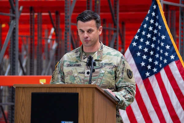 A man wearing a camouflage military uniform stands inside a warehouse behind a podium speaking.