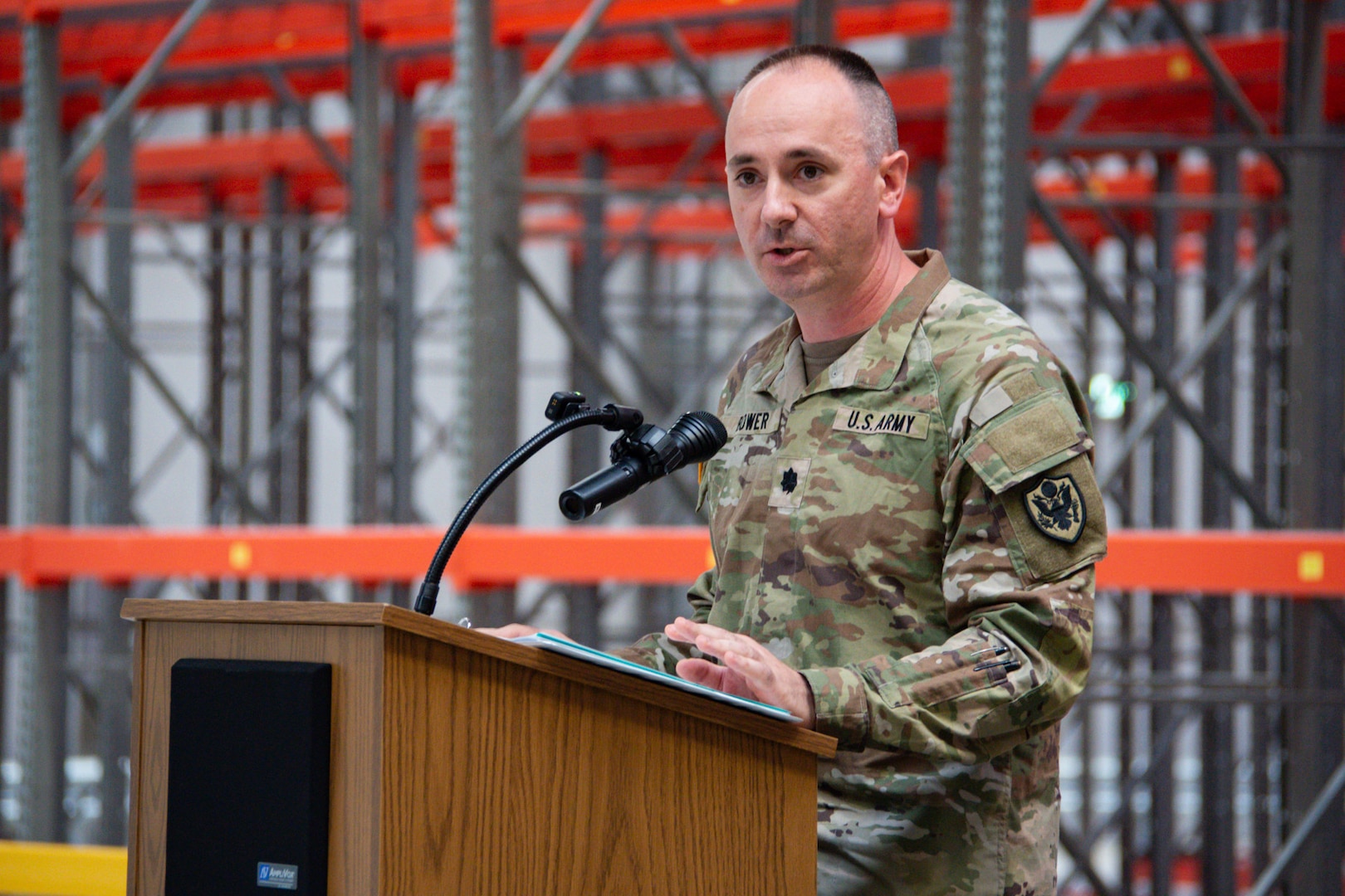 A man wearing a camouflage military uniform stands inside a warehouse behind a podium speaking.