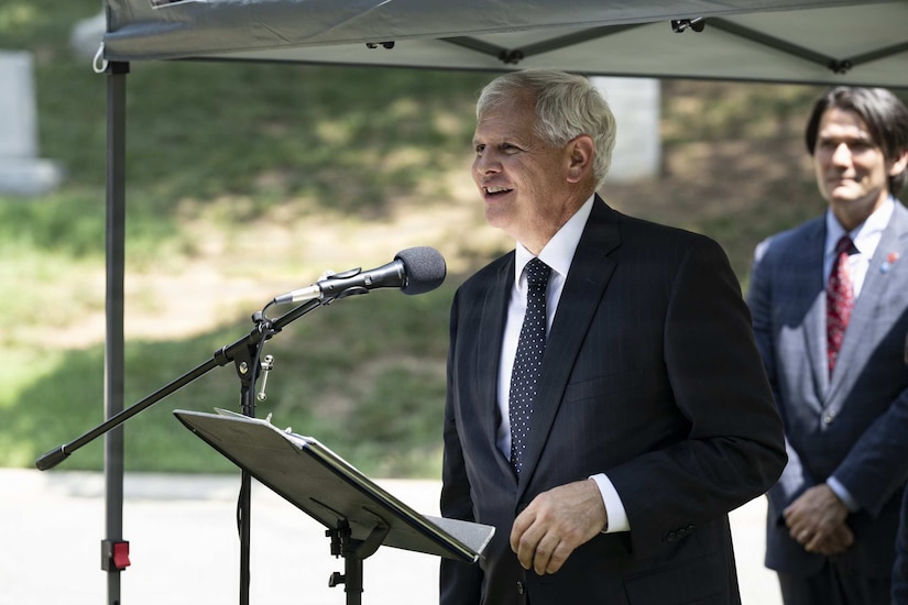 A man in a suit speaks into a microphone during a ceremony outside.