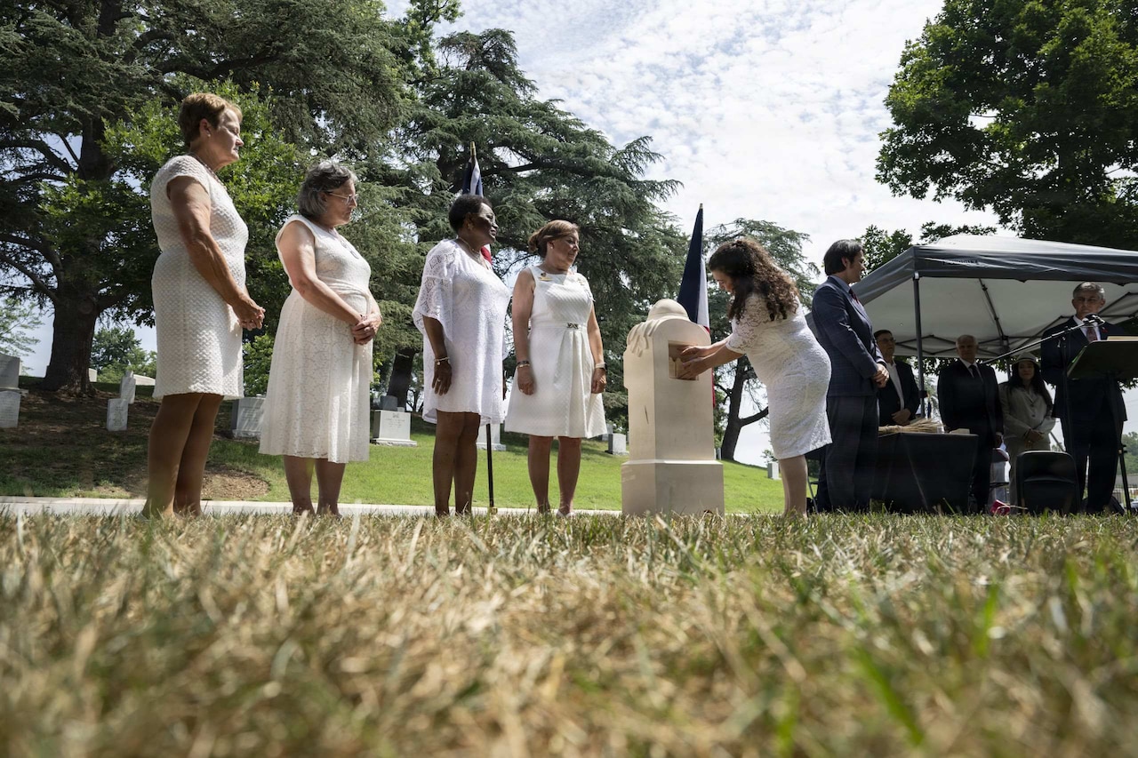 A woman in white formal attire places a container of soil inside a monument at a cemetery while four other women in similar attire stand to the side.