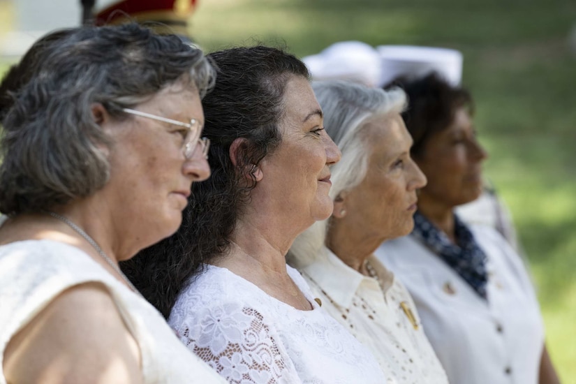 Four women dressed in white formal attire stand outside during a ceremony.