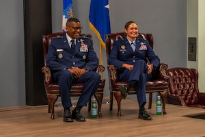 Two military officers in formal uniforms smile while sitting in chairs on a stage.