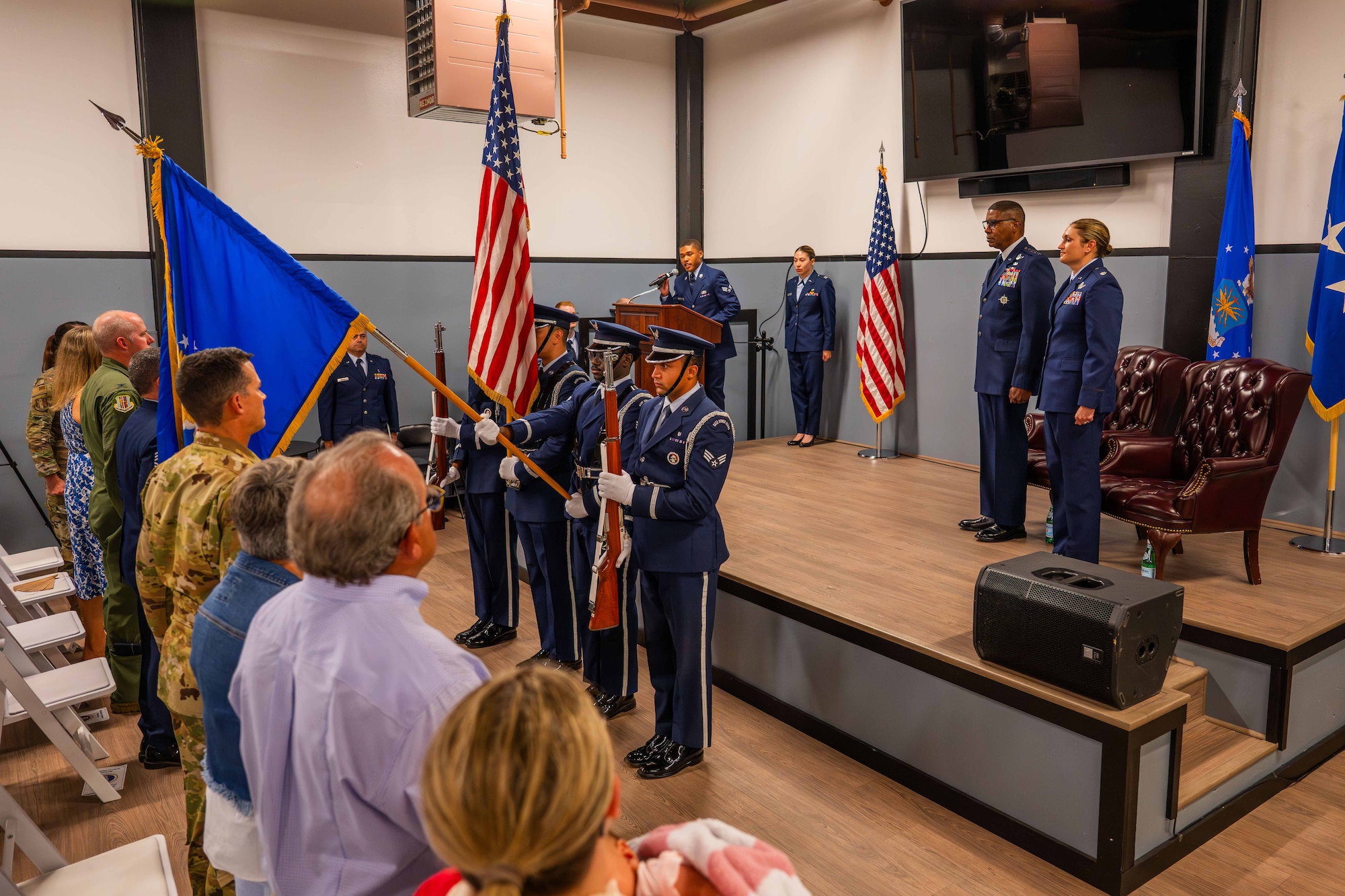 A military  honor guard team presents the colors in front of a crowd during a ceremony inside.