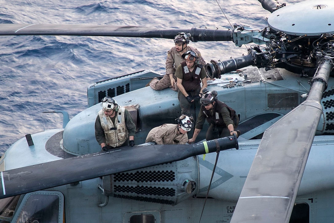 Five Marines work to attach a rotor blade to a helicopter parked on the deck of a ship with water visible in the background during daytime.