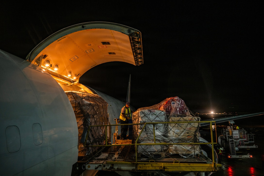 A man wearing a safety vest removes a large pallet of supplies from the side door of a large, parked aircraft at night.