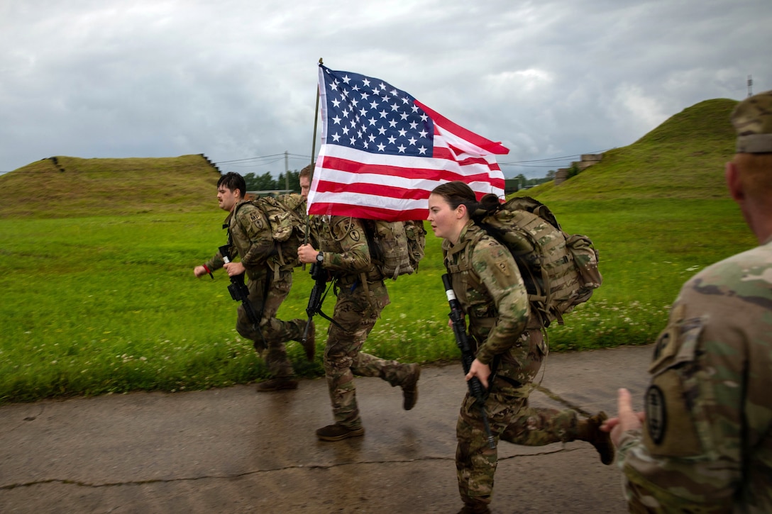 Three soldiers wearing camouflage uniforms and rucksacks run side by side while carrying rifles, with the middle soldier also carrying an American flag, while another soldier wearing a camouflage uniform looks at them and claps his hands.