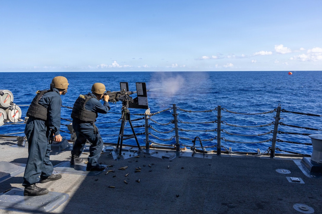 A sailor in camouflage uniform fires a machine gun off the deck of a ship at a floating orange target while another sailor, also in camouflage uniform, observes.