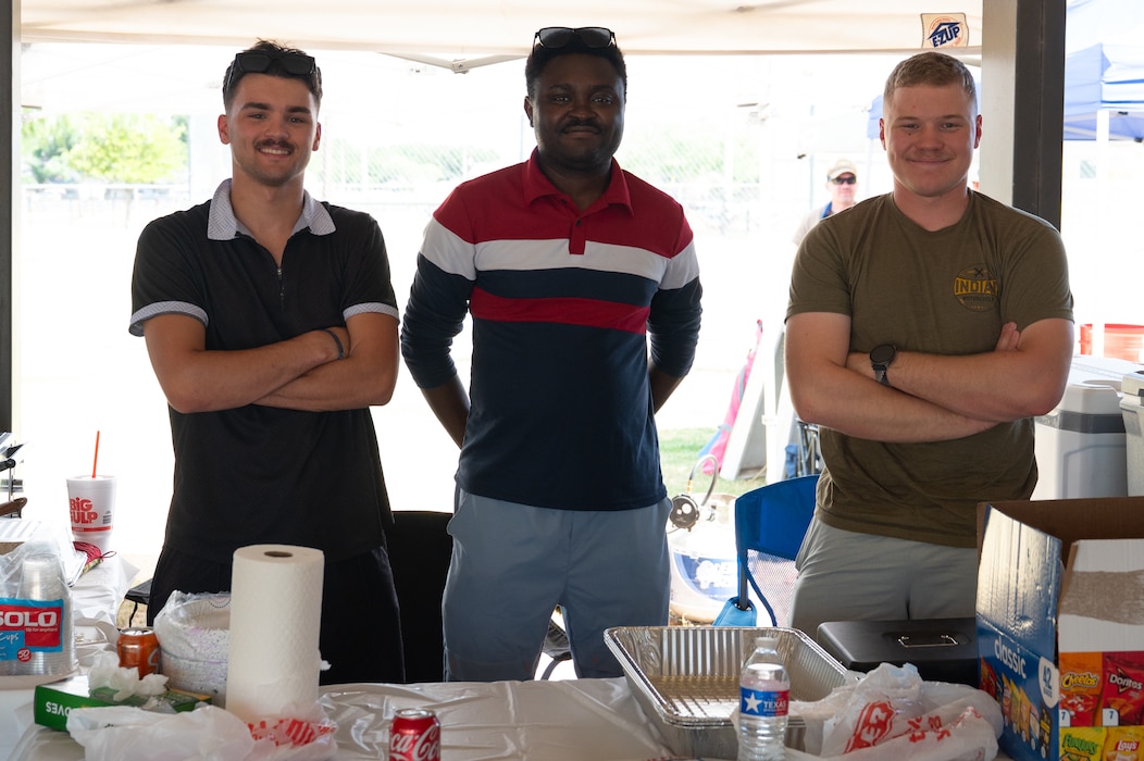 Three individuals in a line are smiling in front of a food spread