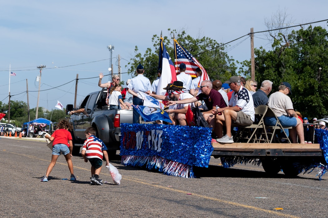 A parade float passes on a road and kids are picking up candy being tossed by participants on the float