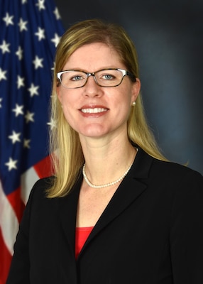 A headshot of a blond woman with glasses in a dark business suit with the American Flag positioned behind her.