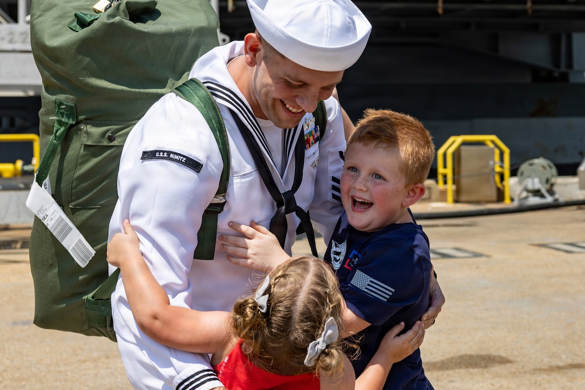 A sailor in a white and blue uniform carrying a green duffle on their back hugs two smiling children with a partially visible boat in the background.