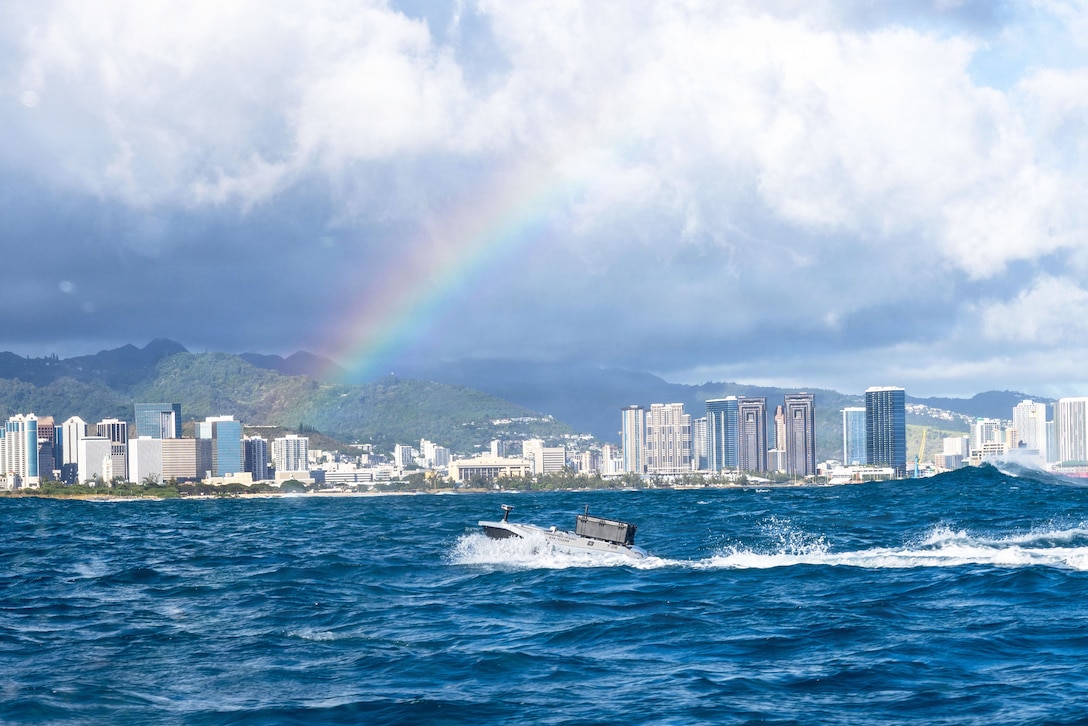 An unmanned surface vessel transits a body of water near a rainbow with an island's skyline and mountains in the background.