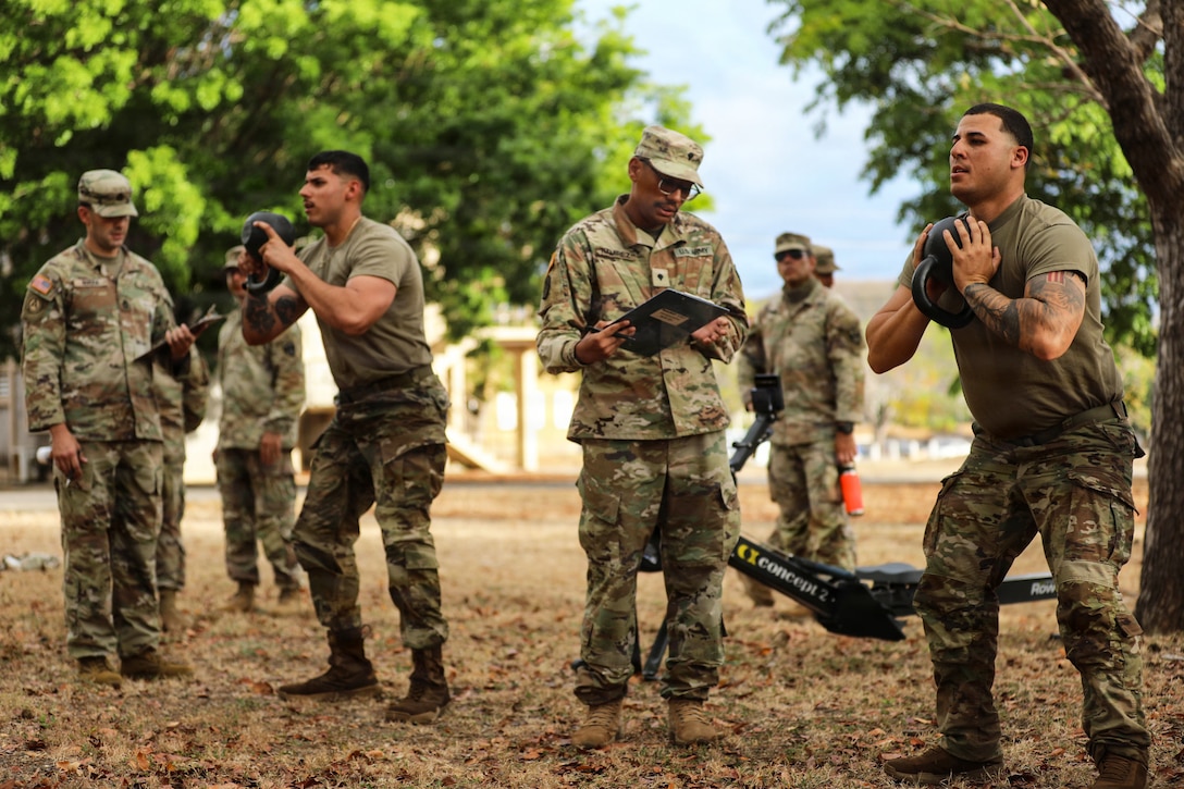 Two soldiers perform squats with kettlebells in a field under trees as soldiers evaluate them with clipboards.