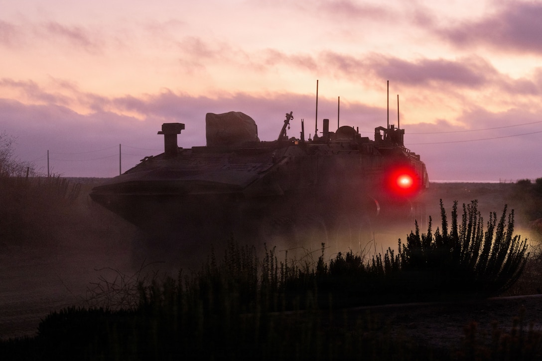 Marines operate an amphibious combat vehicle in a dark field illuminated by a red headlight under a light pinkish cloudy sky.