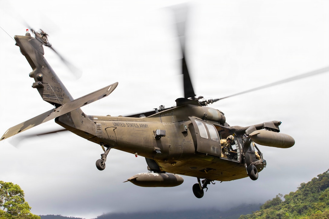 A soldier looks out the open door of an Army helicopter in flight. A gray sky is in the background.