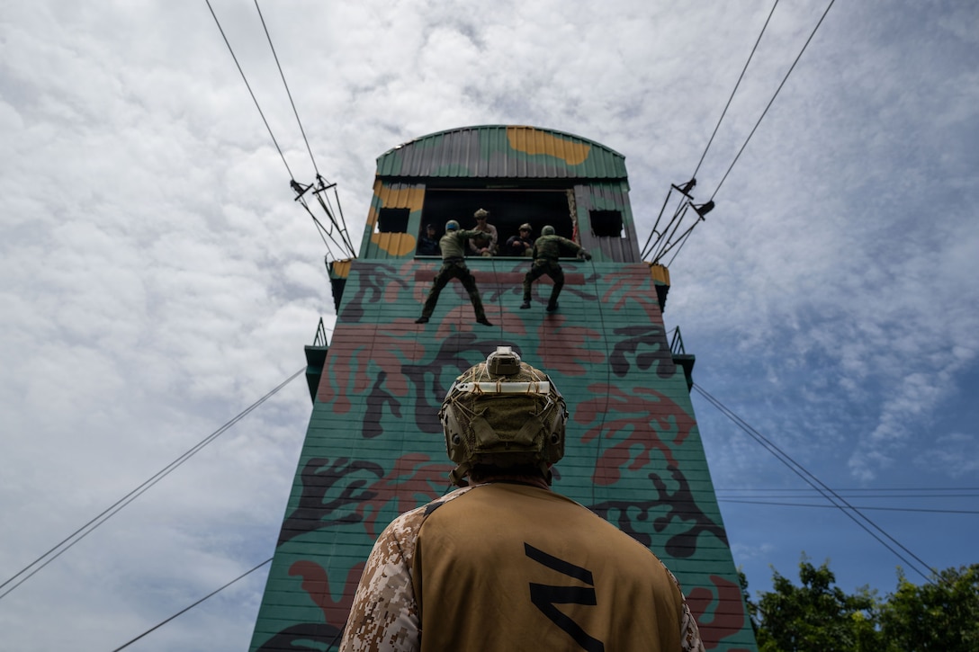 A sailor in a camouflage uniform looks up at two people using ropes to descend a small green tower as other people look down from a tower window.