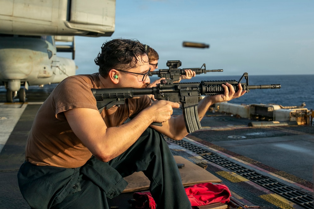 A sailor fires a rifle while crouching on the deck of a ship. Another sailor is doing the same nearby. The horizon and a small black airborne object are in the background.