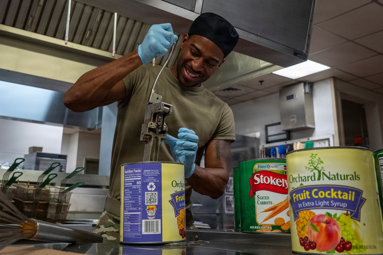 A man wearing a camouflage military uniform uses a large can opener to open two large cans of fruit cocktail and a large can of carrots.
