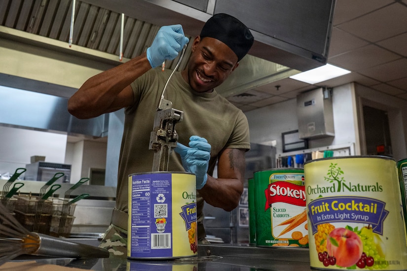 A man wearing a camouflage military uniform uses a large can opener to open two large cans of fruit cocktail and a large can of carrots.