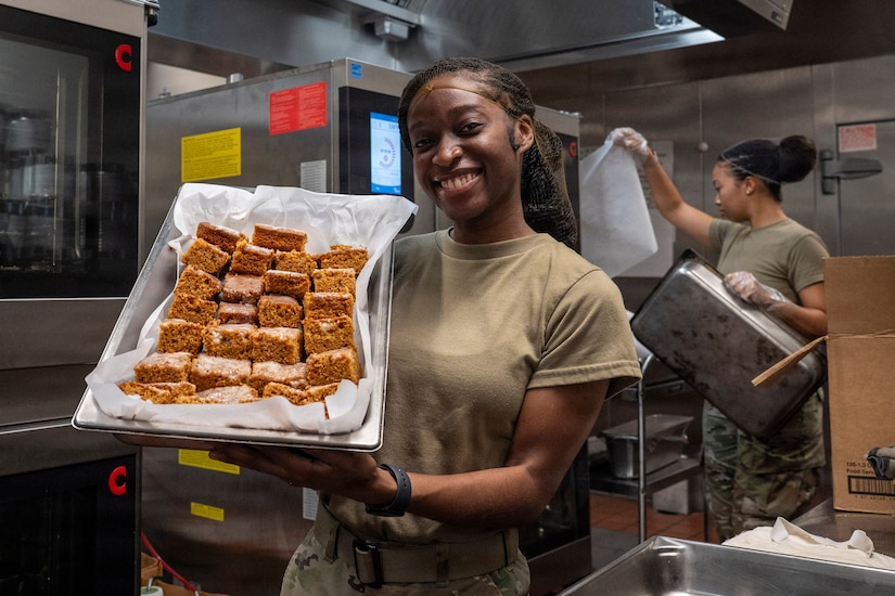 A woman wearing a camouflage military uniform smiles as she holds up a pan of muffins.