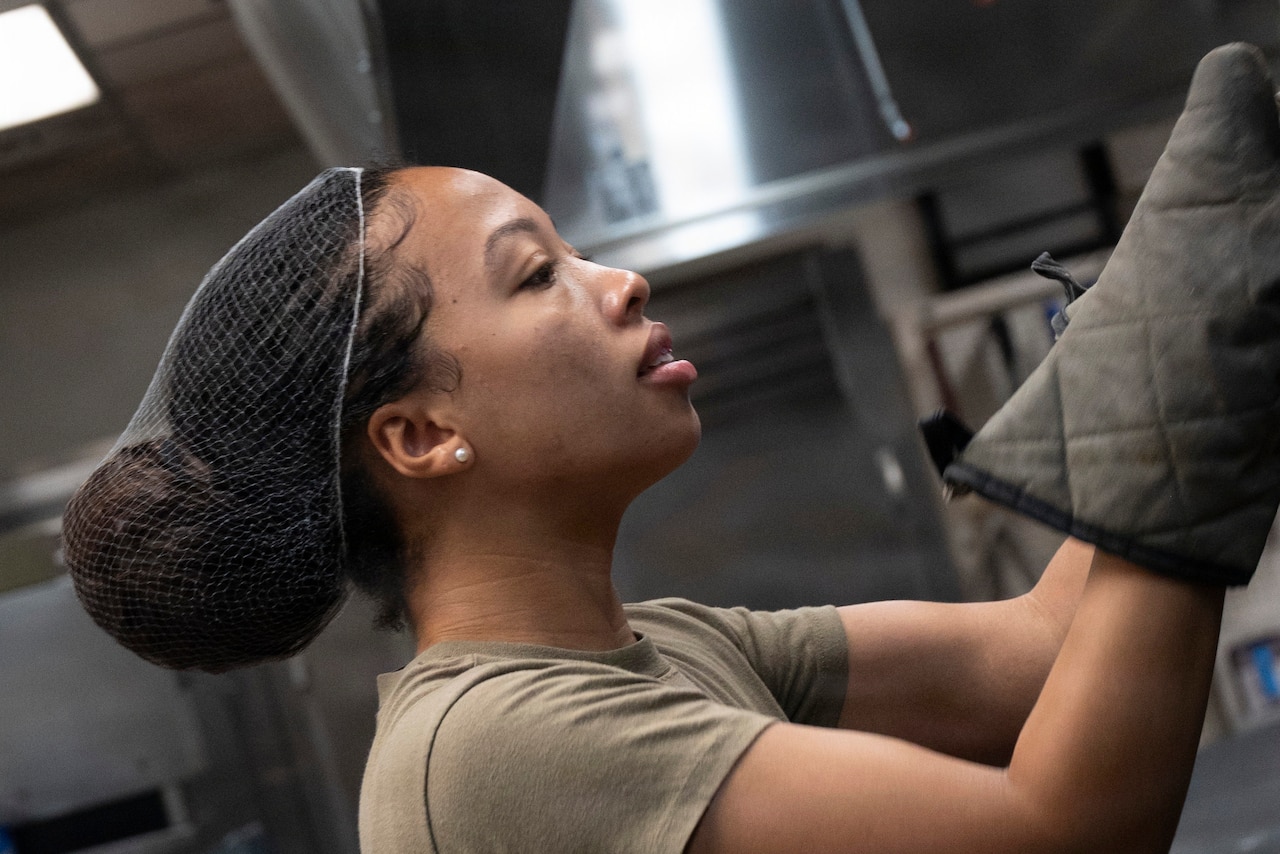 A woman in a camouflage military uniform wears oven mitts as she looks into an oven.