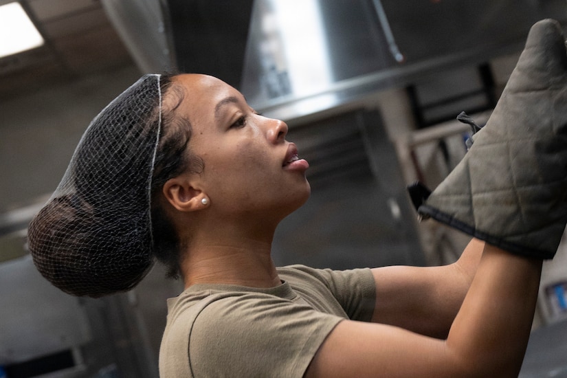A woman in a camouflage military uniform wears oven mitts as she looks into an oven.