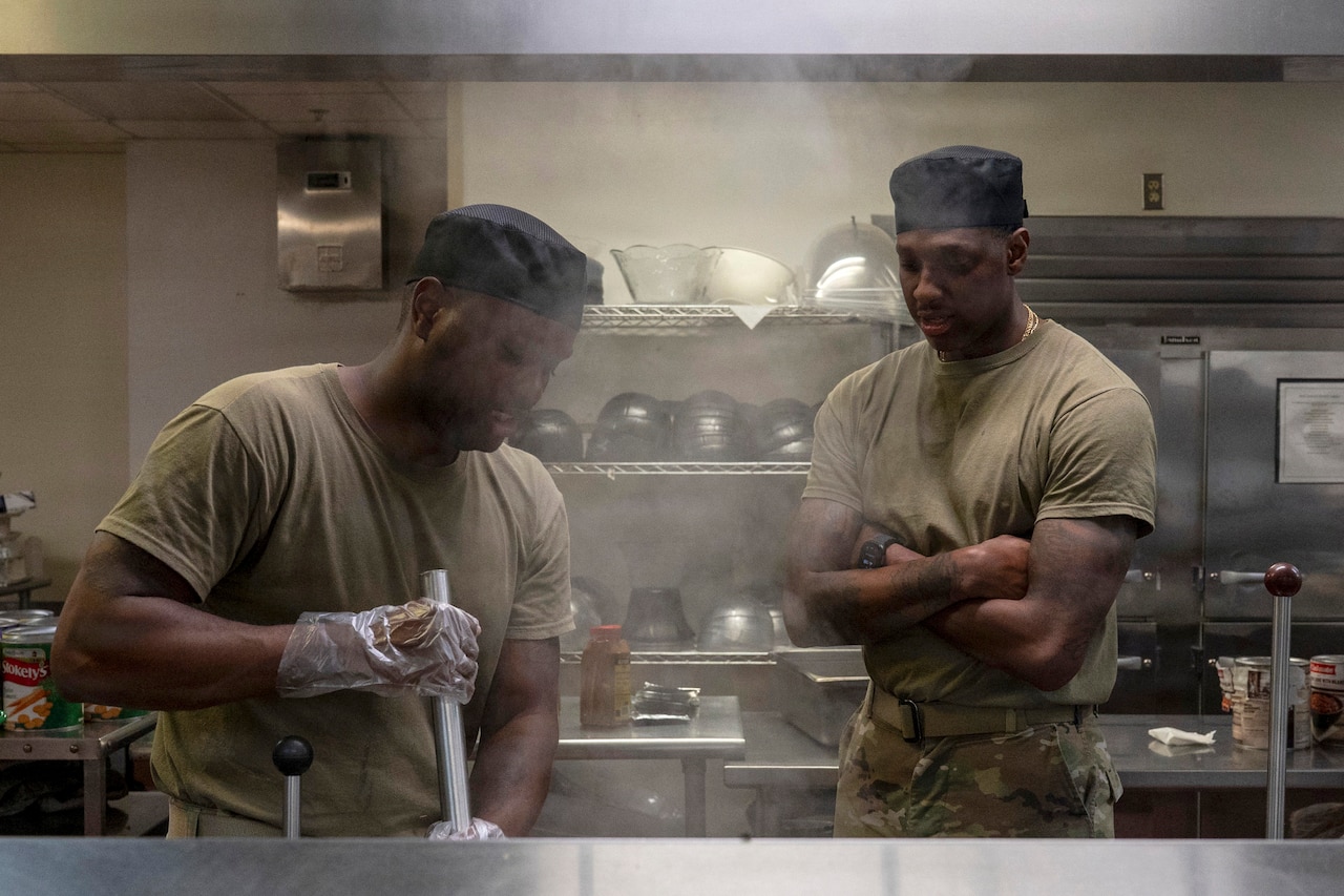 A man wearing a camouflage military uniform stirs soup in a large pot as another man in similar attire looks on.