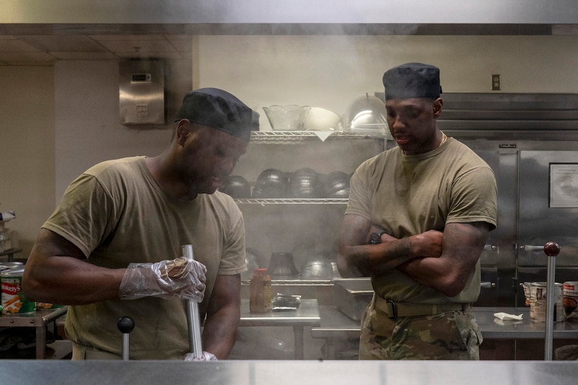 A man wearing a camouflage military uniform stirs soup in a large pot as another man in similar attire looks on.