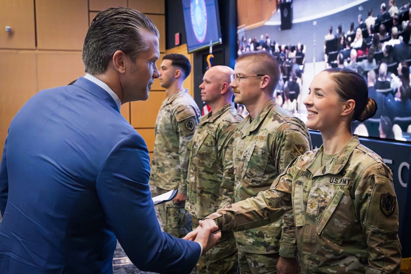 A man in business attire shakes hands with a woman in a camouflage military uniform while three people stand alongside her in similar attire.