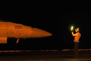 A sailor in yellow shirt and helmet gestures toward a jet using a light baton on a ship deck in the dark.