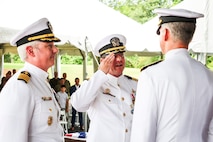 Photograph showing three naval officers in white dress uniforms engaged in a formal ceremony outdoors wiith white tents in the background. One officer is saluting another, with visible medals and insignia on their uniforms, while people observe in the background.
