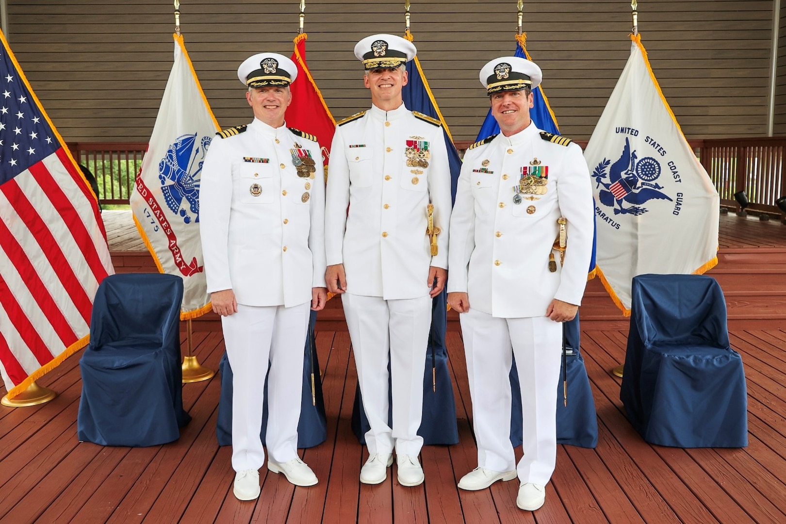 Photograph of three U.S. Navy officers in white dress uniforms standing on a wooden deck with six flags behind them, including the American flag and various military branch flags. The officers display multiple medals and insignias, indicating rank and achievements, with five empty blue-covered chairs behind them and front of the flags.