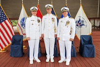 Photograph of three U.S. Navy officers in white dress uniforms standing on a wooden deck with six flags behind them, including the American flag and various military branch flags. The officers display multiple medals and insignias, indicating rank and achievements, with five empty blue-covered chairs behind them and front of the flags.