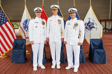 Photograph of three U.S. Navy officers in white dress uniforms standing on a wooden deck with six flags behind them, including the American flag and various military branch flags. The officers display multiple medals and insignias, indicating rank and achievements, with five empty blue-covered chairs behind them and front of the flags.