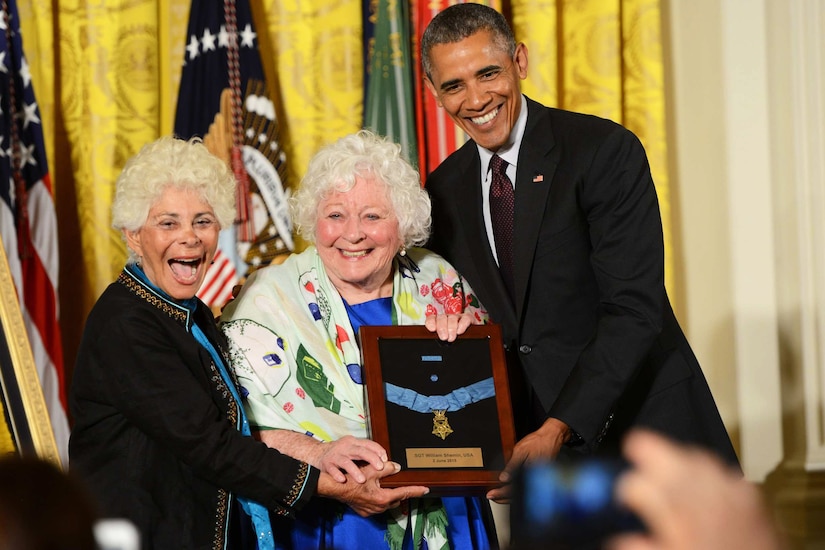Two elderly women and a man smile while holding a framed medal with a ribbon.
