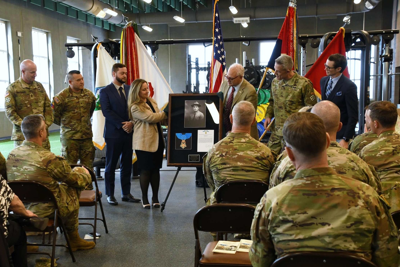 A man and a woman in business attire unveil a plaque in a large room with other people in similar attire and camouflage military uniforms standing on the side and sitting in chairs.
