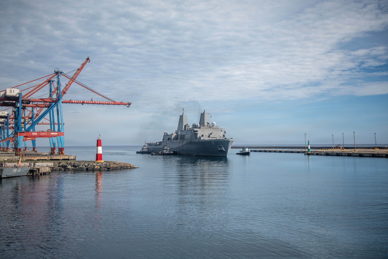 A large military ship sails in a harbor while being guided by three tugboats.