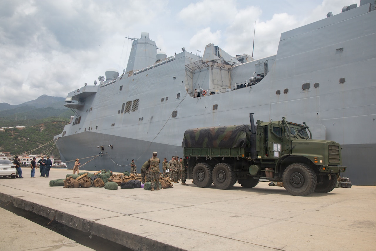 A small group of people in camouflage military uniforms take equipment off the back of a military vehicle parked on a pier; there is a large ship docked in the background.