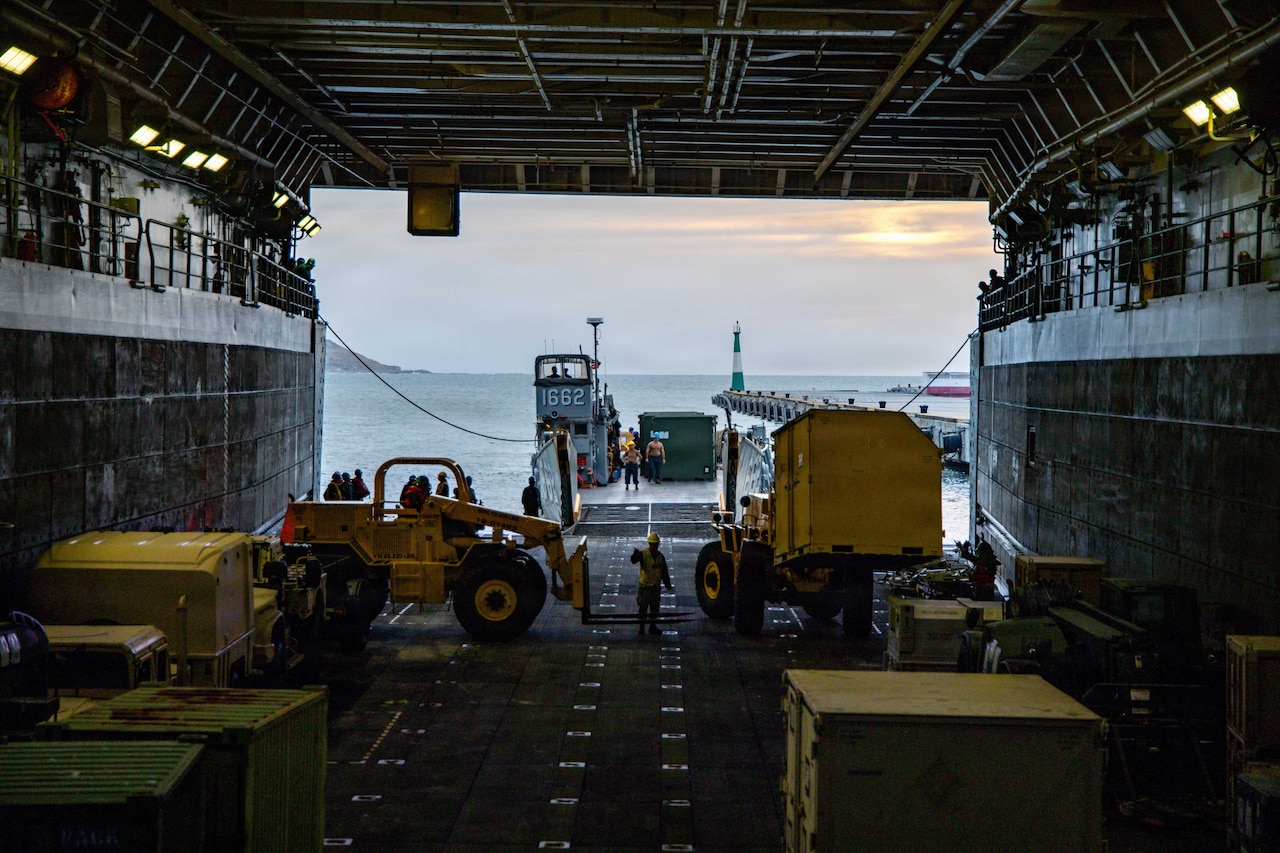 A dozen people in military uniforms and hard hats use forklifts to transport containers from the cargo bay of a large military ship onto another transport dock ship.