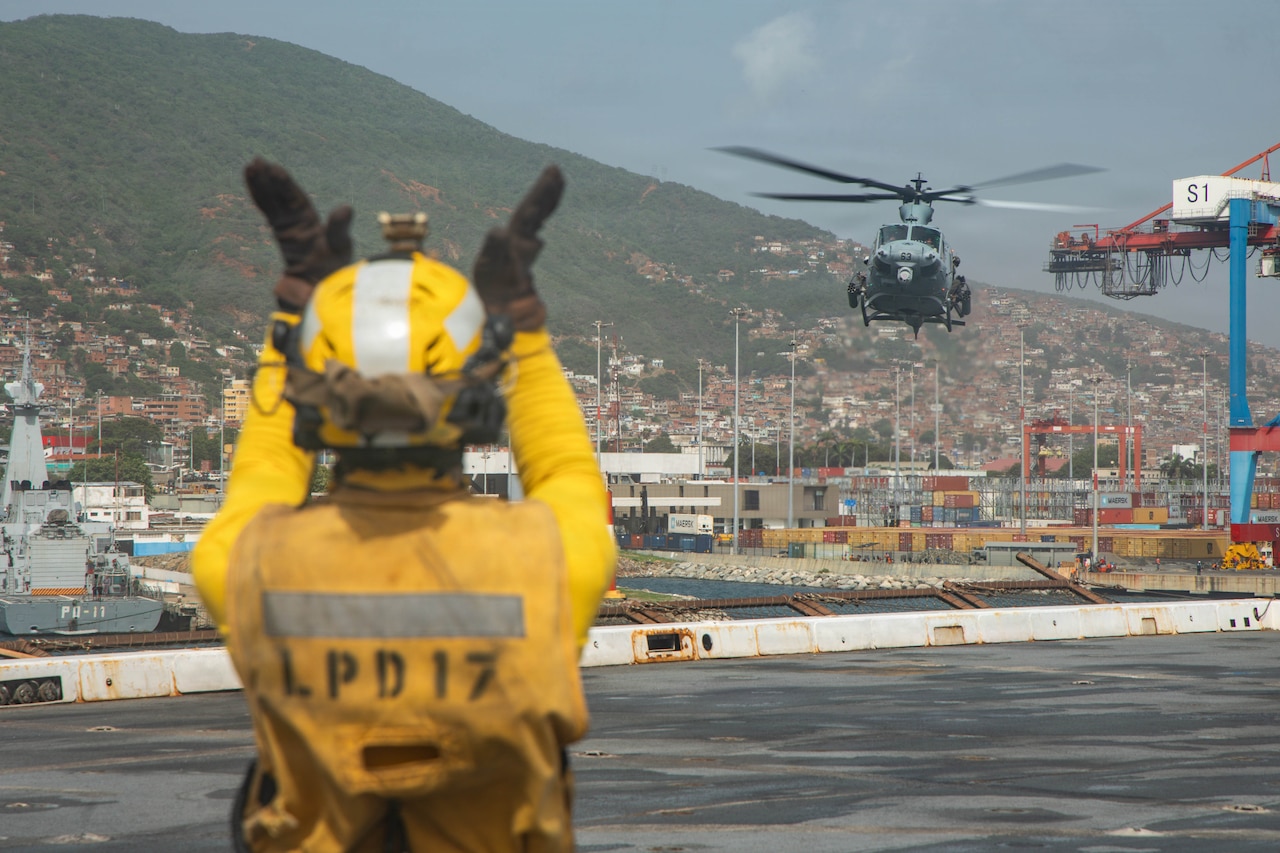 A person in a military flight deck uniform gives hand signals to helicopter pilots as they prepare to land on the deck of a ship in the water.