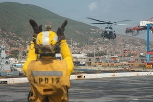 A person in a military flight deck uniform gives hand signals to helicopter pilots as they prepare to land on the deck of a ship in the water.