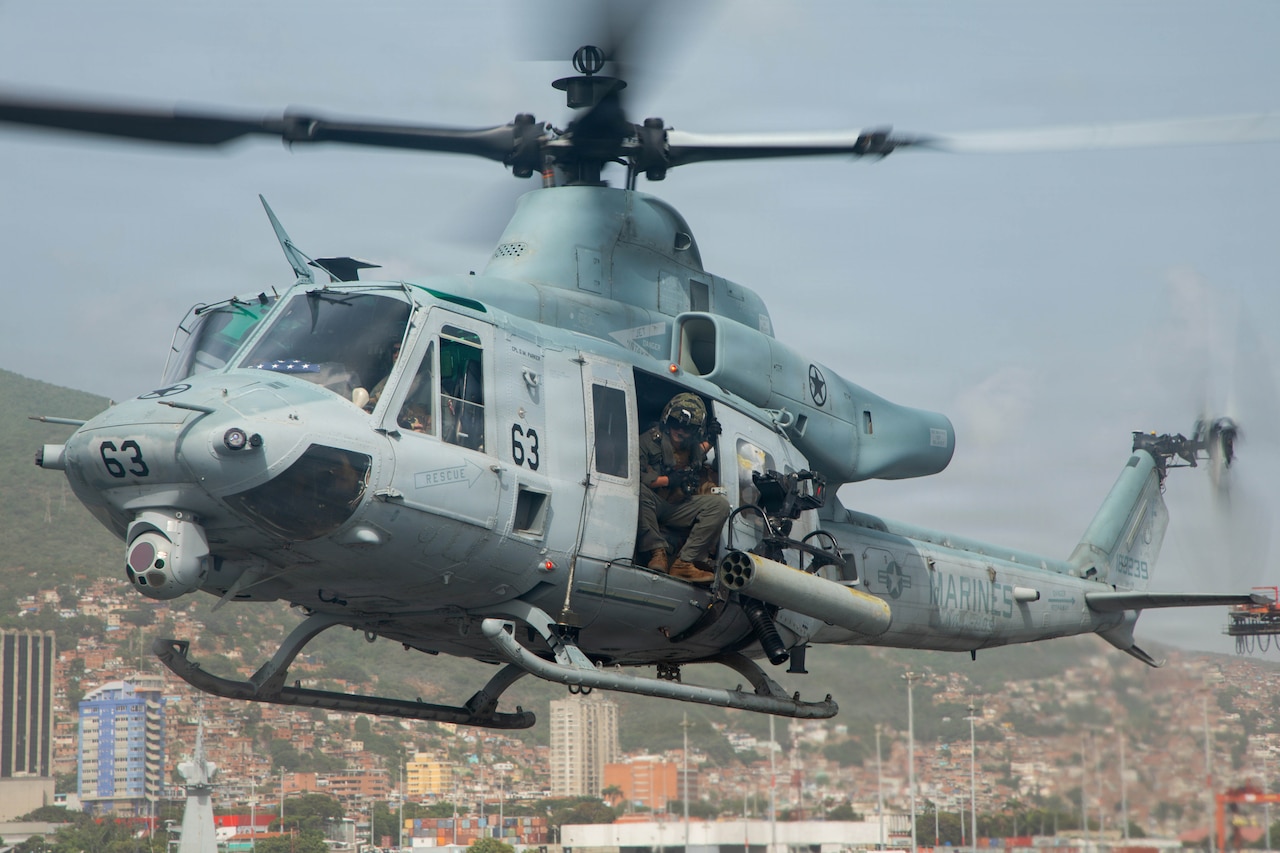 A person in a flight suit looks out the door of a military helicopter as it prepares to land on the deck of a ship in the water.